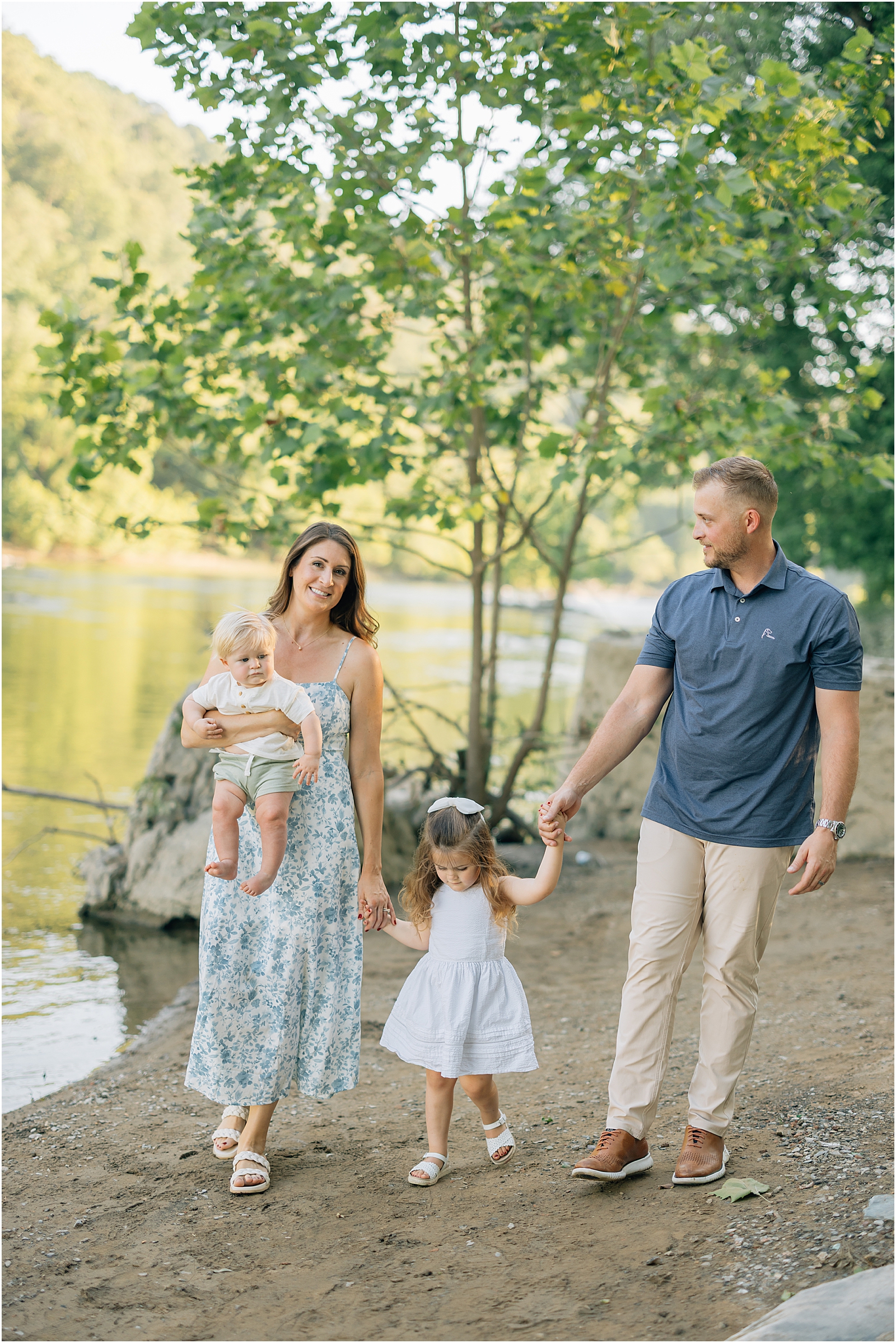 family of four holds hands walking on riverside bank in historic harpers ferry