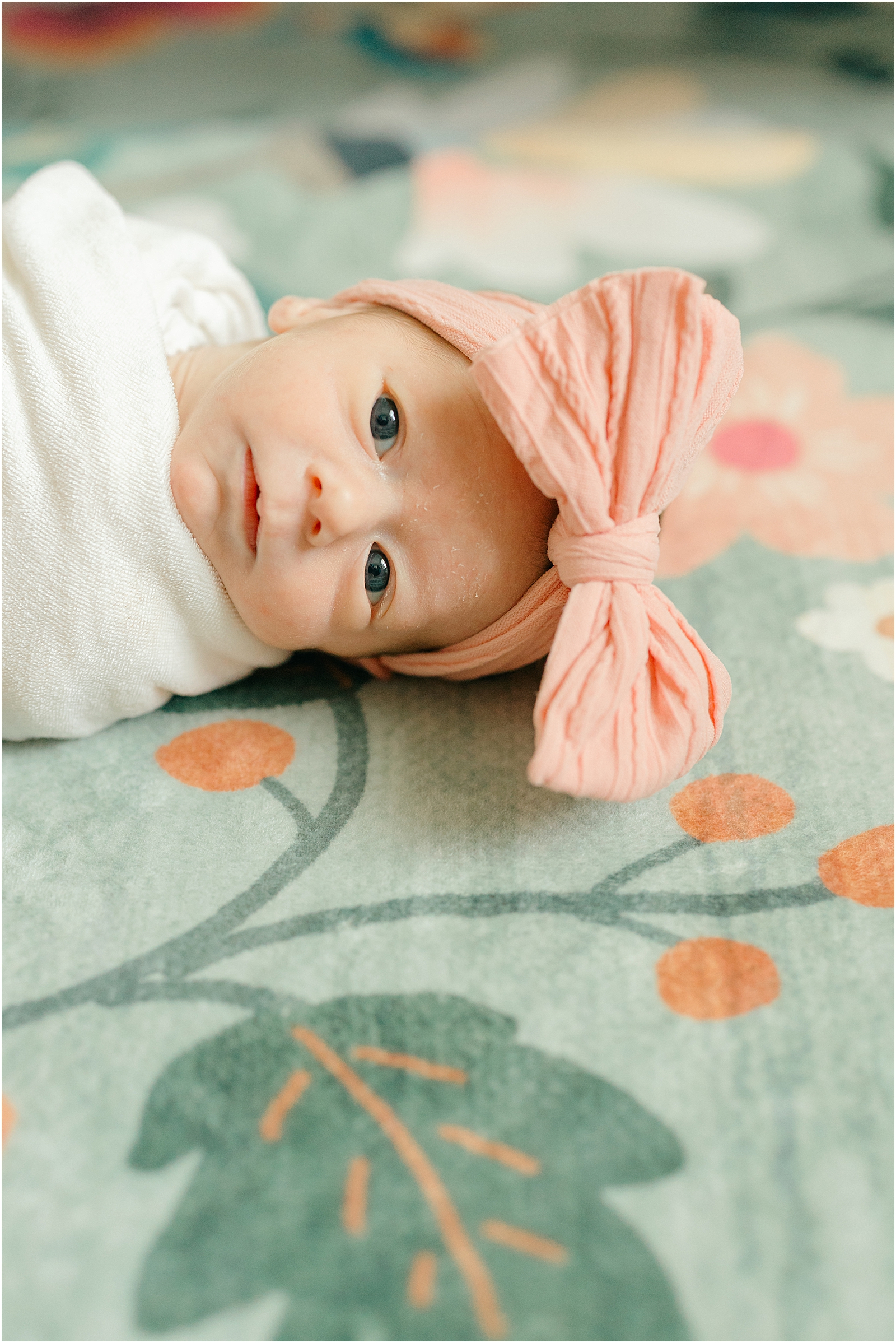 newborn girl wearing pink bow swaddled in white blanket laying on colorful nursery rug